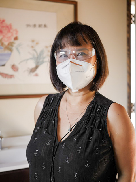 Lynn Cheng, acupuncturist and herbalist, standing in front of a Chinese painting with a KN-95 mask and protective glasses on